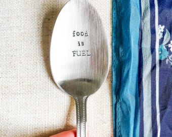 May include: A silver spoon with the stamped text "food is FUEL". The spoon has a decorative handle and is set against a white and blue background. A unique kitchen utensil for serving.