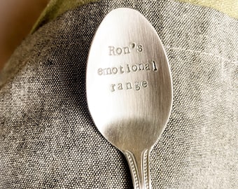 May include: A silver spoon with the words "Ron's emotional range" stamped on the spoon's bowl. The spoon is resting on a grey and green textured fabric.