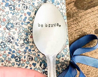 May include: A silver spoon with the stamped text "be brave," held over a floral patterned fabric and a blue ribbon. The spoon has a decorative handle and is set against a wooden surface.