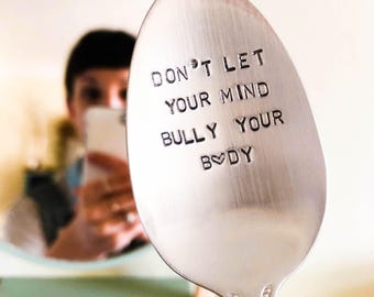 May include: A stainless steel spoon with the words "DON'T LET YOUR MIND BULLY YOUR BODY" stamped on the spoon's surface. The spoon has a heart symbol in place of the letter "O" in the word "BODY".