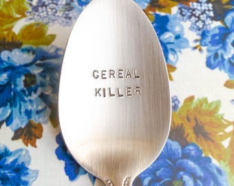 May include: A silver-toned metal spoon with a brushed finish featuring the stamped text CEREAL KILLER centred on the bowl. The spoon has a decorative beaded pattern along the handle neck, providing a clear view of the engraved lettering.