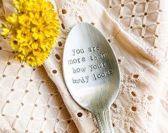 May include: A silver spoon with the stamped text "You are more than how your body looks" rests on a cream-coloured, embroidered cloth. A cluster of yellow flowers is in the upper left corner.