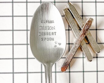 May include: A silver dessert spoon stamped with the words "Custom DESIGN DESSERT SPOON" on the bowl, with a star, flower, and heart below. Several metal stamps are next to the spoon. The spoon is on a checkered surface.