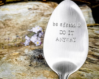 May include: A silver spoon with the words "be afraid & DO IT ANYWAY" stamped on the bowl. The spoon is set against a rustic wooden background with small purple flowers.