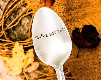 May include: A silver spoon with the text "YOU'VE GOT THIS" stamped on it. The spoon is set against a backdrop of autumn leaves and a textured, golden-brown fabric. A yellow leaf and a red leaf are also visible.