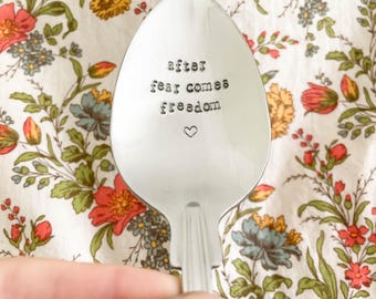 May include: A silver spoon with the stamped text "after fear comes freedom" and a small heart symbol. The spoon is set against a floral patterned background with red, yellow, and blue flowers.