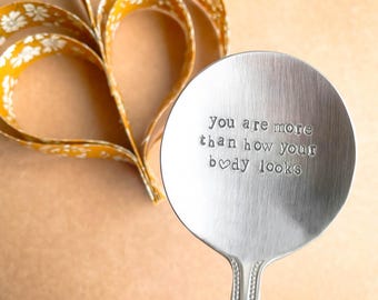 May include: A silver spoon with the stamped text "You are more than how your body looks." A yellow floral ribbon is in the background, shaped into a heart. The spoon has a rounded bowl and a decorative handle.