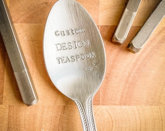 May include: A silver teaspoon with the words "Custom DESIGN TEASPOON" stamped on the bowl, alongside a star, heart, and flower. The spoon rests on a wooden surface, with other metal tools visible.