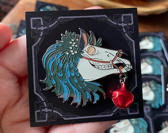 May include: A metal enamel pin featuring a white horse skull with a teal and green mane, adorned with a floral crown. The skull wears a red bridle and has a small, dangling red jingle bell attached to the bit. The pin is mounted on a black card.