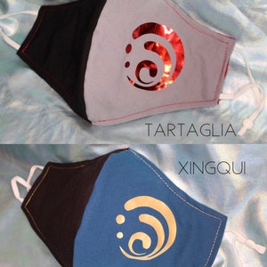 Pode incluir: Duas m&aacute;scaras faciais com designs diferentes. A m&aacute;scara superior &eacute; cinza com um design de redemoinho vermelho e o texto "TARTAGLIA". A m&aacute;scara inferior &eacute; azul com um design de redemoinho dourado e o texto "XINGQIU".