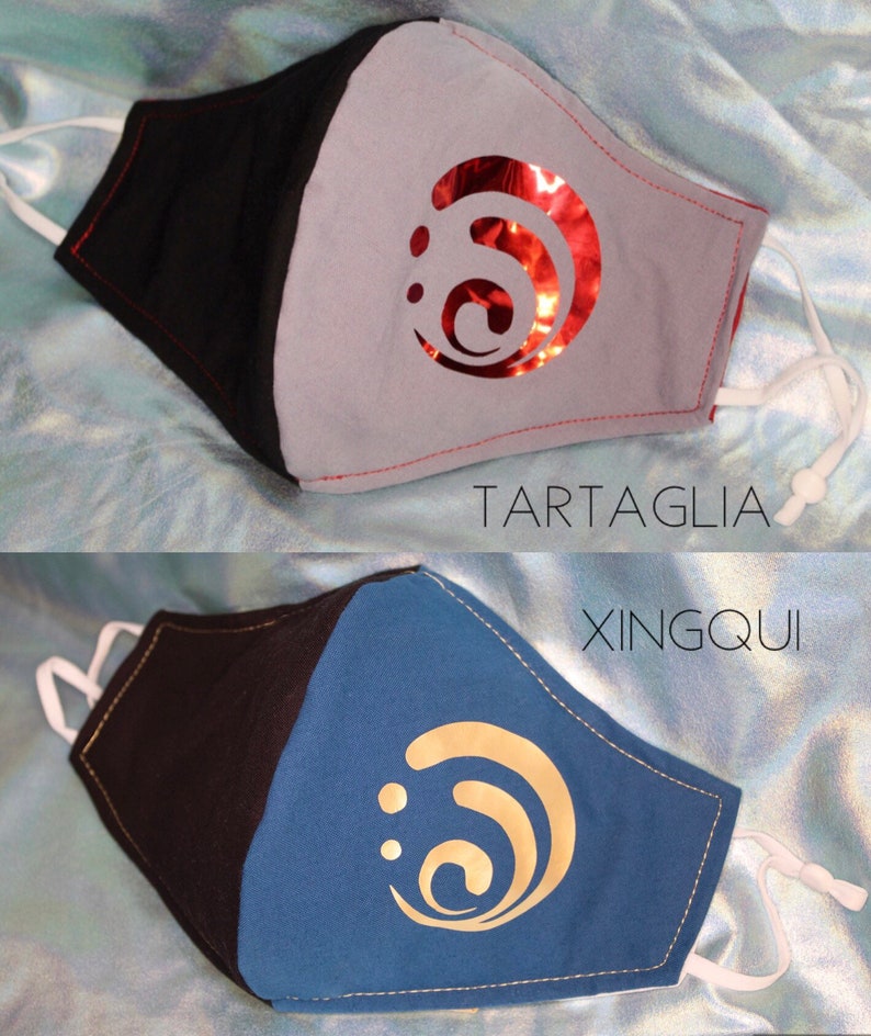 Pode incluir: Duas m&aacute;scaras faciais com designs diferentes. A m&aacute;scara superior &eacute; cinza com um design de redemoinho vermelho e o texto "TARTAGLIA". A m&aacute;scara inferior &eacute; azul com um design de redemoinho dourado e o texto "XINGQIU".