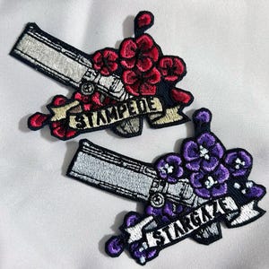 Könnte beinhalten: Zwei gestickte Bügelbilder mit einer Pistole und Blumen. Der erste Patch ist rot und weiß mit dem Text "STAMPEDE". Der zweite Patch ist lila und weiß mit dem Text "STARGAZE".