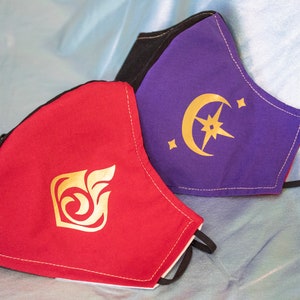 Pode incluir: Duas m&aacute;scaras faciais, uma vermelha com um design de espiral dourado e outra roxa com um design de lua crescente e estrela douradas.