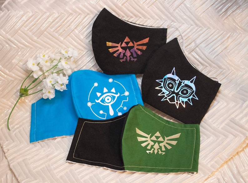 Op de afbeelding: Vijf stoffen mondmaskers met verschillende ontwerpen uit de videogame-serie The Legend of Zelda. De maskers zijn zwart, blauw, groen en twee zijn zwart met verschillende ontwerpen. De ontwerpen zijn de Triforce, het Sheikah-oog, het Gerudo-symbool en het Majora's Mask.