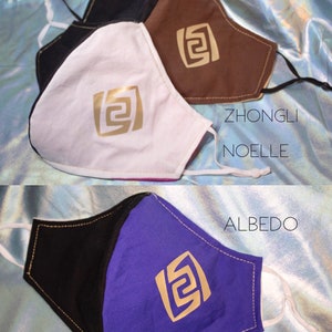 Pode incluir: Tr&ecirc;s m&aacute;scaras faciais de tecido preto, branco, marrom e roxo. Cada m&aacute;scara tem um design geom&eacute;trico dourado e os nomes "Zhongli", "Noelle" e "Albedo" escritos nelas.
