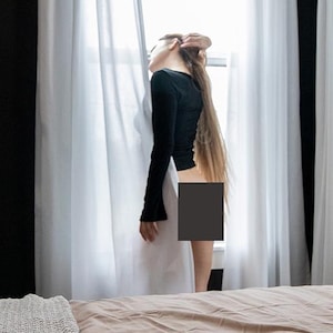 May include: A bedroom scene with a bed covered in a tan comforter and a white knitted throw blanket. A black bookshelf displays decorative items. White curtains frame a window, and a gold lamp stands nearby.