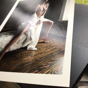 May include: A rectangular photographic print featuring a person with dark skin posing on a dark wooden floor. The person is draped in a white fabric and positioned near a textured, light-coloured fur-like material against a dark background.