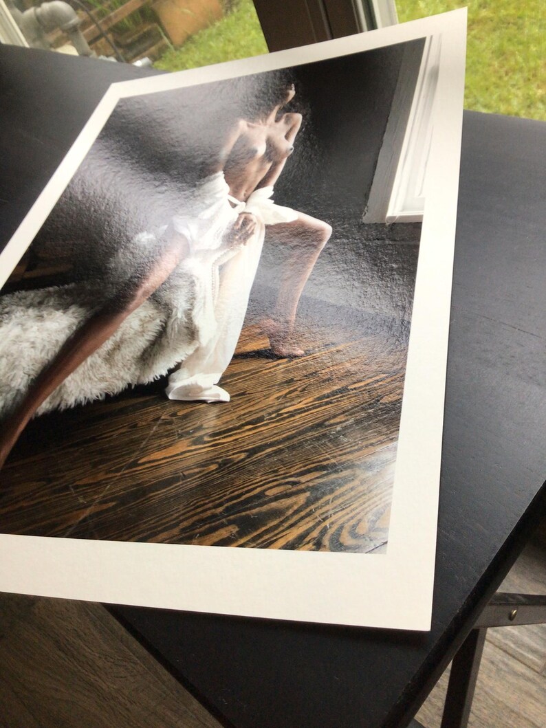 May include: A rectangular photographic print featuring a person with dark skin posing on a dark wooden floor. The person is draped in a white fabric and positioned near a textured, light-coloured fur-like material against a dark background.