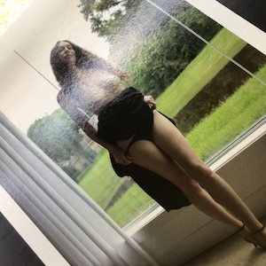 May include: A photographic print featuring a person with long dark hair standing in front of a window. The person is wearing a black skirt and black undergarments, with the fabric of the skirt held slightly to the side, revealing the legs.