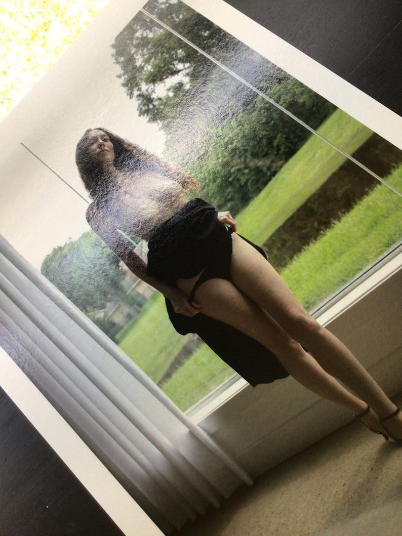 May include: A photographic print featuring a person with long dark hair standing in front of a window. The person is wearing a black skirt and black undergarments, with the fabric of the skirt held slightly to the side, revealing the legs.