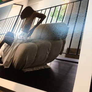 May include: A photographic print featuring a person with long dark hair sitting on a light-colored, multi-sectioned upholstered chair. The scene includes a black metal railing in the background and dark wooden flooring, presented within a white border.