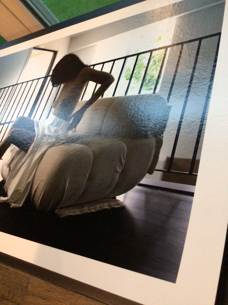 May include: A photographic print featuring a person with long dark hair sitting on a light-colored, multi-sectioned upholstered chair. The scene includes a black metal railing in the background and dark wooden flooring, presented within a white border.