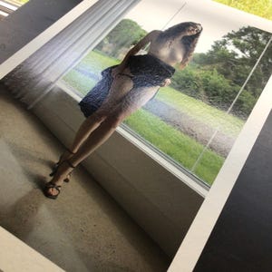 May include: A photographic print featuring a person with long dark hair standing in front of a window. The person is wearing a dark blue textured skirt and black strappy high-heeled sandals. The scene shows a green outdoor landscape visible through the glass.