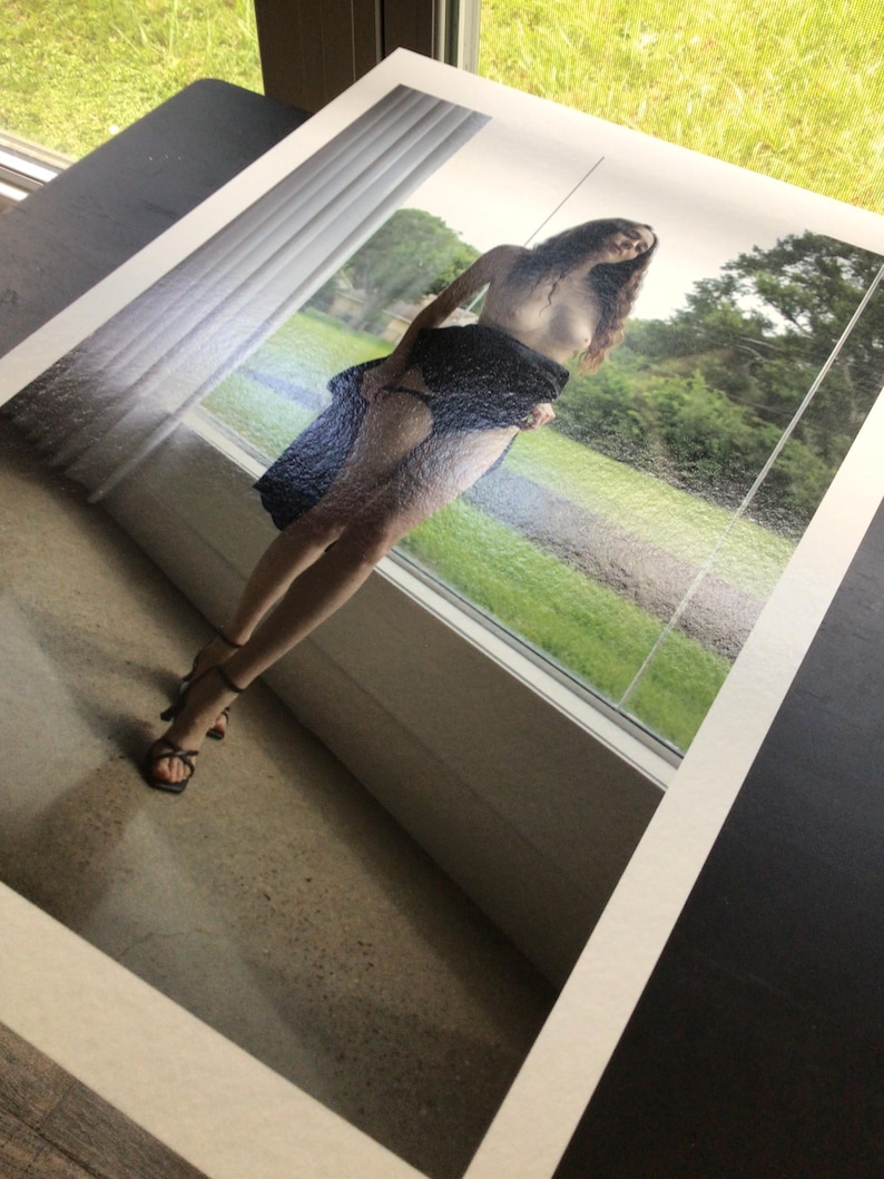 May include: A photographic print featuring a person with long dark hair standing in front of a window. The person is wearing a dark blue textured skirt and black strappy high-heeled sandals. The scene shows a green outdoor landscape visible through the glass.