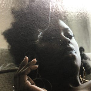 May include: A person with dark skin exhales smoke, with the smoke curling upwards. The person is wearing large hoop earrings. A lit cigarette is held in their hand. The image is a close-up.