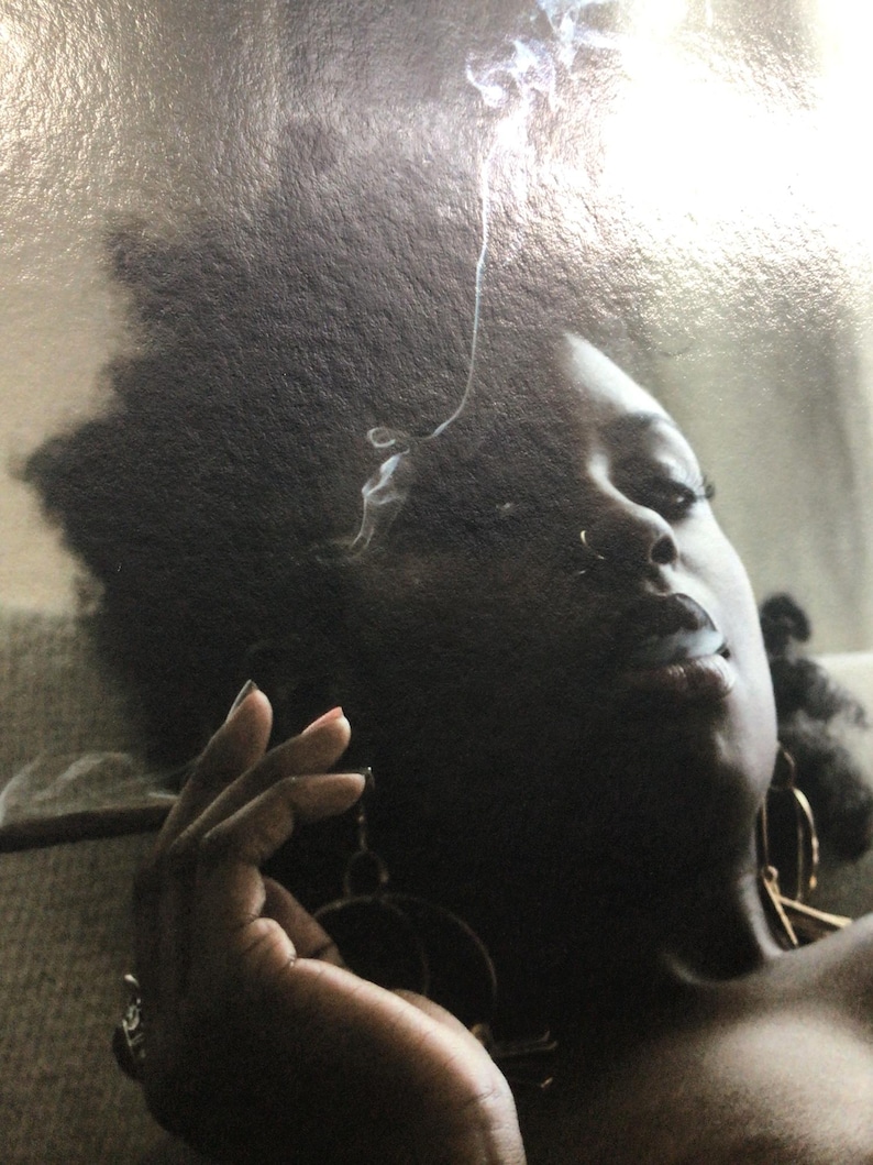 May include: A person with dark skin exhales smoke, with the smoke curling upwards. The person is wearing large hoop earrings. A lit cigarette is held in their hand. The image is a close-up.