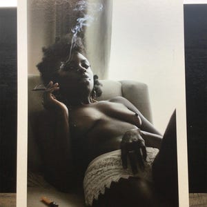 May include: A black and white photograph of a person reclining on a chair, smoking. The person is wearing a white lace garment. Smoke rises from the cigarette. A lighter is on the surface.