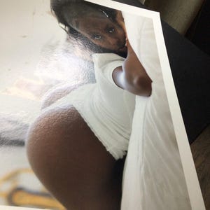 May include: A rectangular photographic print featuring a high-angle view of a person with dark skin wearing a white short-sleeved shirt, positioned against a white surface, with a blurred yellow object visible in the lower left corner of the print.