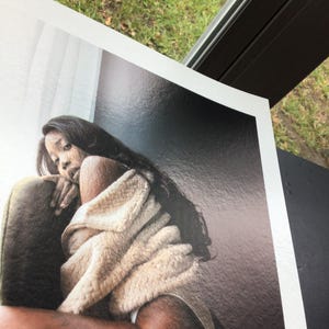 May include: A print of a person with long dark hair, wrapped in a beige blanket, sitting in a green chair. The print has a glossy finish and is set against a dark background.