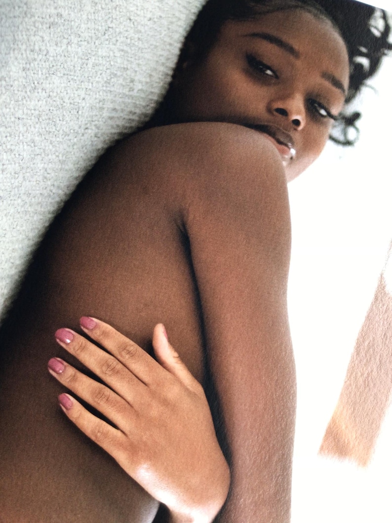 May include: A person with dark skin and long eyelashes looks over their shoulder. Their hand, with pink nail polish, is placed on their chest. The person is lying on a white surface.