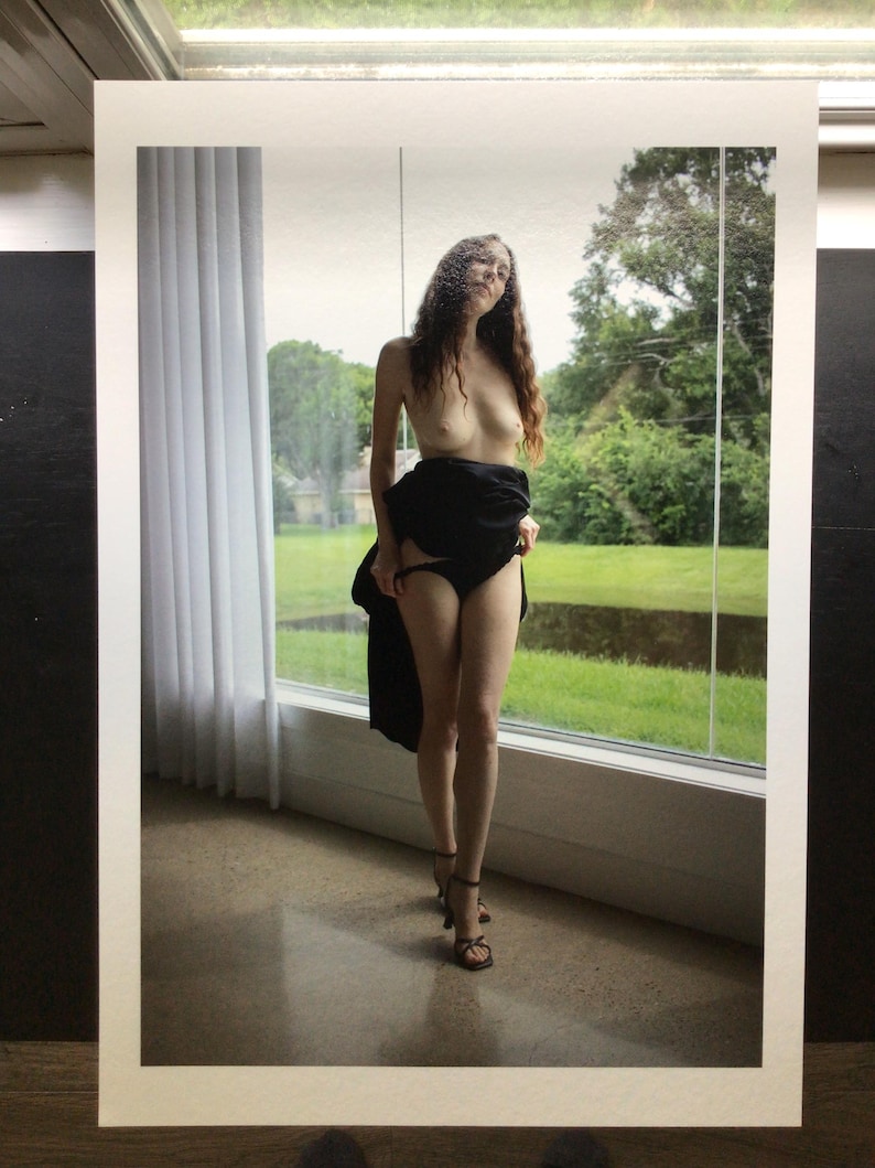 May include: A photographic print featuring a person with long wavy hair standing in front of a large window. The person is wearing black underwear and a black garment draped around the waist, paired with black strappy high-heeled sandals.
