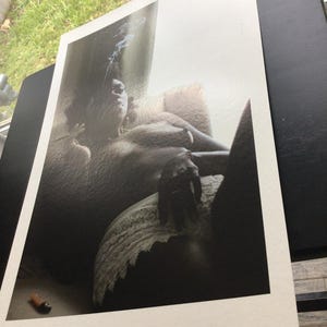 May include: A monochrome art print showing a person reclining, with smoke rising. The print is on white paper, with a lighter and a cigarette visible in the foreground. The image is displayed on a black surface.