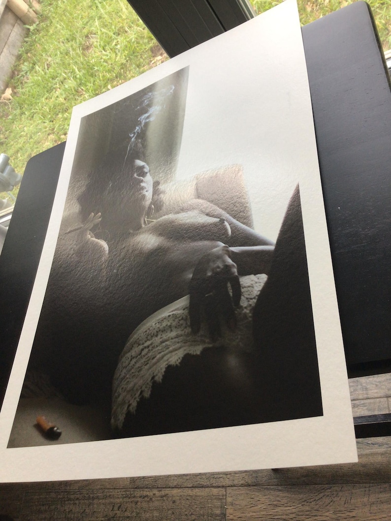 May include: A monochrome art print showing a person reclining, with smoke rising. The print is on white paper, with a lighter and a cigarette visible in the foreground. The image is displayed on a black surface.