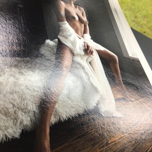 May include: A photographic print featuring a person with dark skin sitting on a white faux fur bench. The person is draped in a sheer white fabric garment that falls loosely around the waist and legs, contrasting against a dark floor background.