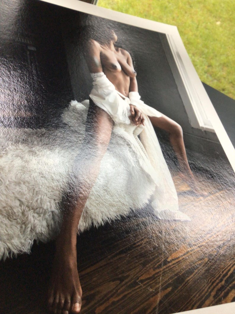 May include: A photographic print featuring a person with dark skin sitting on a white faux fur bench. The person is draped in a sheer white fabric garment that falls loosely around the waist and legs, contrasting against a dark floor background.