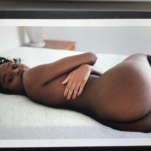 May include: A person with dark skin lies on a white surface, arms crossed. The person's gaze is directed towards the viewer. The image is a portrait, with soft lighting and a focus on the subject's form.
