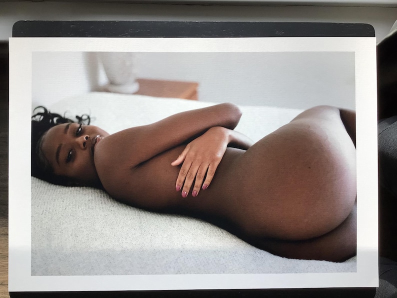 May include: A person with dark skin lies on a white surface, arms crossed. The person's gaze is directed towards the viewer. The image is a portrait, with soft lighting and a focus on the subject's form.