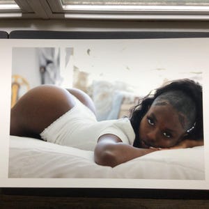 May include: A photographic print featuring a person with dark skin and long dark hair lying on a white surface. The person is wearing a white short-sleeved bodysuit with a ruffled edge and is looking directly toward the camera with a neutral expression.