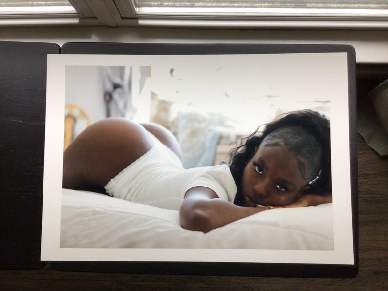May include: A photographic print featuring a person with dark skin and long dark hair lying on a white surface. The person is wearing a white short-sleeved bodysuit with a ruffled edge and is looking directly toward the camera with a neutral expression.