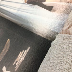 May include: Close-up of a textured, dark surface with a light-colored design and text. A light-colored knitted blanket is in the lower right corner. The image has a soft, muted color palette.