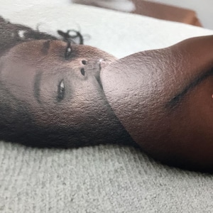 May include: A close-up photograph of a person's face and shoulder. The person has dark skin and is lying on a light gray, textured surface. The lighting is soft, and the focus is on the person's face.