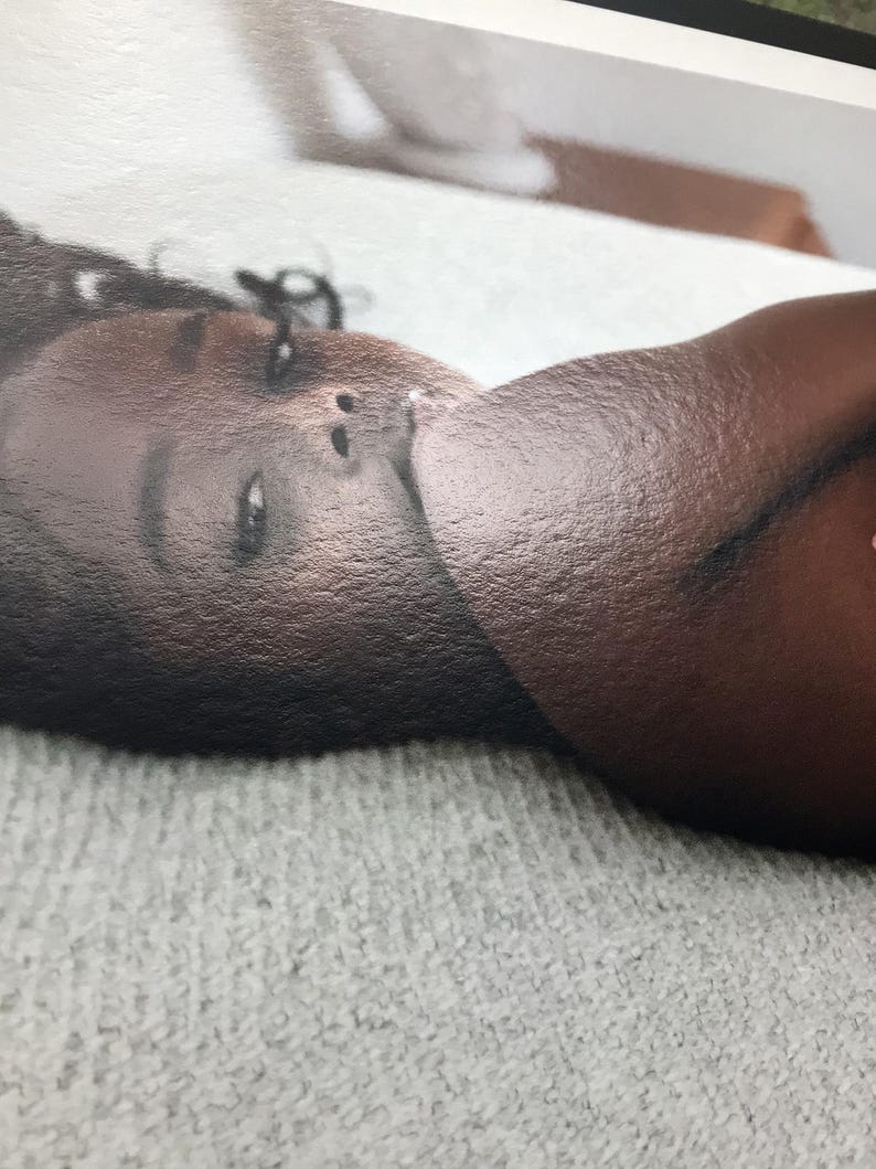 May include: A close-up photograph of a person's face and shoulder. The person has dark skin and is lying on a light gray, textured surface. The lighting is soft, and the focus is on the person's face.