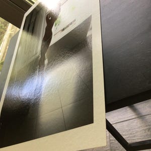 K&ouml;nnte beinhalten: Ein rechteckiger Fotoabzug mit wei&szlig;em Rand, der ein kontrastreiches Bild einer Silhouette in einem hellen Raum mit Fliesenboden zeigt. Der Abzug hat eine gl&auml;nzende Oberfl&auml;che und ist in einem Winkel positioniert.