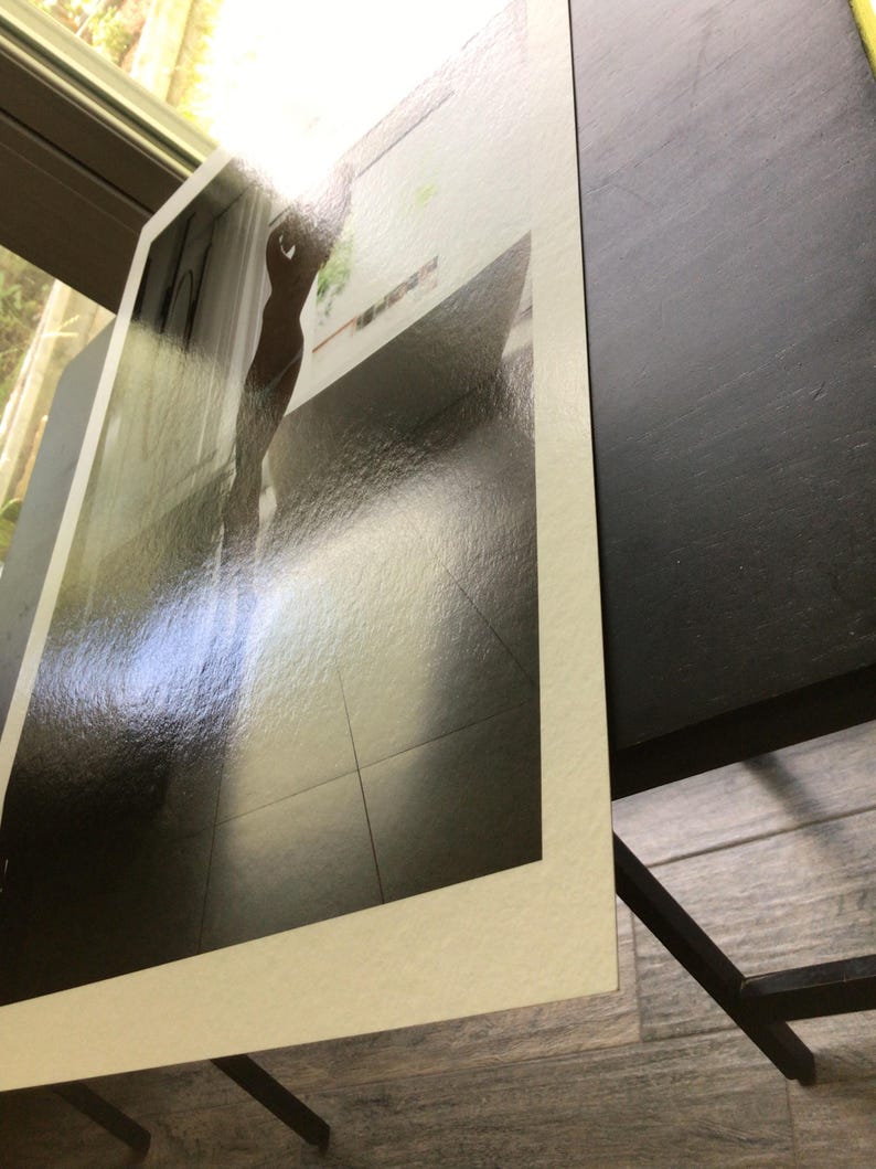 K&ouml;nnte beinhalten: Ein rechteckiger Fotoabzug mit wei&szlig;em Rand, der ein kontrastreiches Bild einer Silhouette in einem hellen Raum mit Fliesenboden zeigt. Der Abzug hat eine gl&auml;nzende Oberfl&auml;che und ist in einem Winkel positioniert.