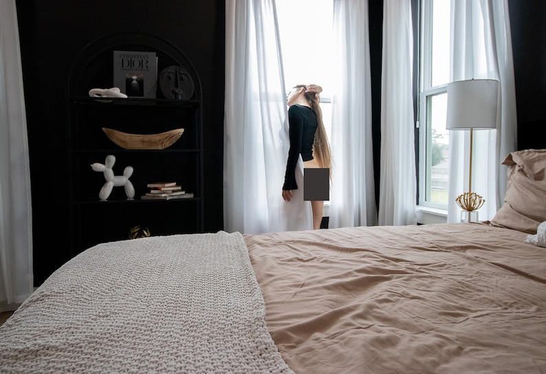 May include: A bedroom scene with a bed covered in a tan comforter and a white knitted throw blanket. A black bookshelf displays decorative items. White curtains frame a window, and a gold lamp stands nearby.