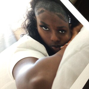May include: A person with dark hair styled in a partial shave on the side, wearing a plain white t-shirt. The individual is resting their head on a white pillow, looking directly toward the camera with a neutral expression and dark eyeliner.